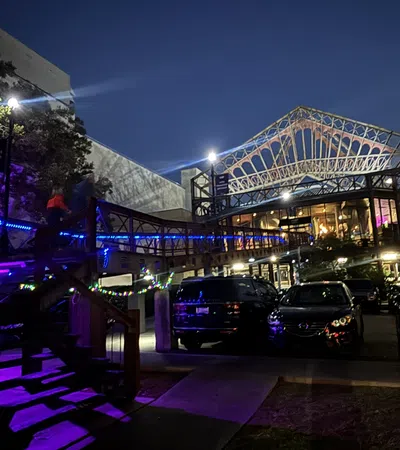 Night view of a modern building with illuminated wooden stairs and parked cars under a steel framework roof structure.