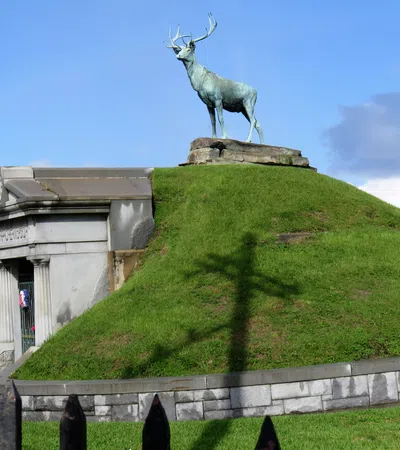 Bronze stag statue atop green grassy mound at historic cemetery under blue sky with scattered clouds.