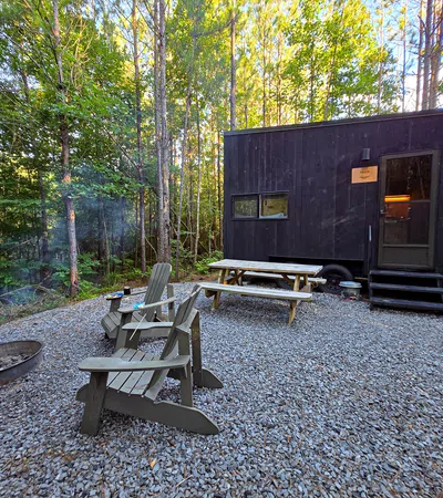Tiny black cabin in forest clearing with picnic table, Adirondack chairs, and a fire pit on gravel ground.