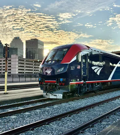 Amtrak train parked on tracks at a station with city skyline and vibrant cloudy sky at sunset.