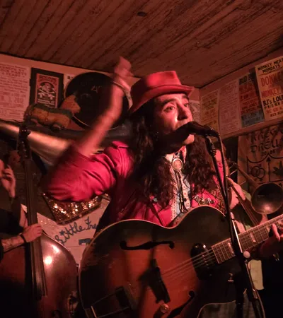 Live band performing in a cozy, dimly lit venue with musician singing and playing guitar surrounded by brass instruments.