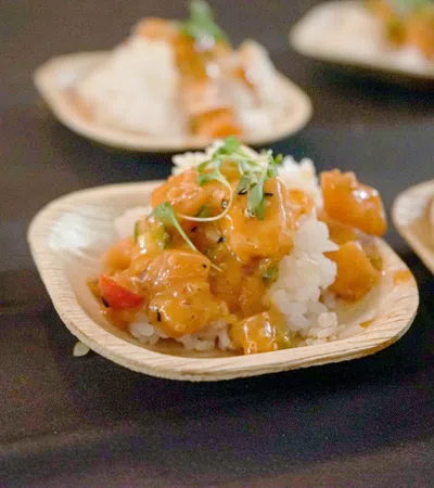 Close-up of seafood poke bowls served on small biodegradable plates with rice and garnished herbs on black table.