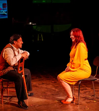 Man and woman in bright clothes sit facing each other on chairs in a dark theater setting with a screen in the background.
