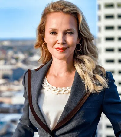 Professional woman with blonde hair smiling confidently in business attire against urban cityscape background.