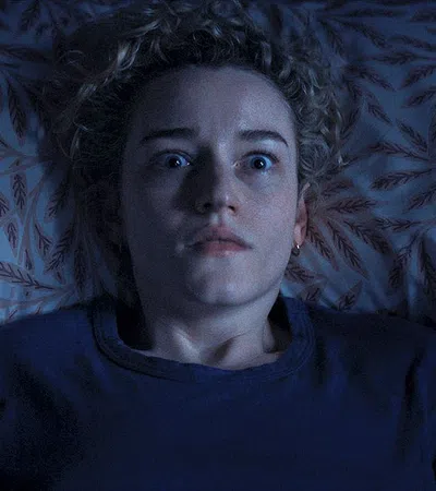 Woman lying in bed staring wide-eyed, appearing shocked or scared in dim, blue-toned lighting.