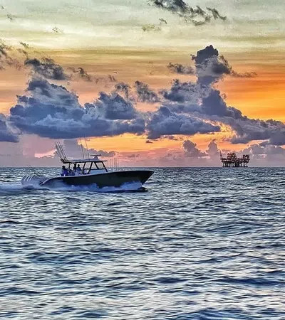 Speedboat cruising on ocean water against a dramatic sunset with scattered clouds and an offshore platform in view.