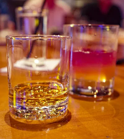 Two glasses on a wooden table, one with whiskey and the other with a layered cocktail, blurred background.