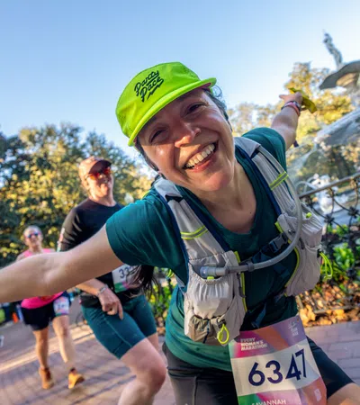 Smiling female marathon runner wearing neon green cap and bib 6347 gestures with arms wide on park path.