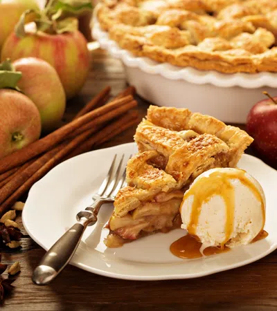 Slice of homemade apple pie with vanilla ice cream and caramel sauce on white plate with fresh apples and cinnamon sticks.