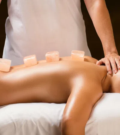 Woman receiving hot stone massage therapy with pink salt stones on her back in a spa setting.