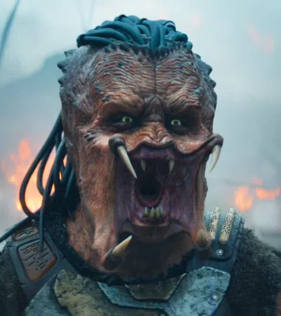 Close-up of a fierce alien creature with sharp teeth, dreadlock-like hair, and armor in smoky battlefield.