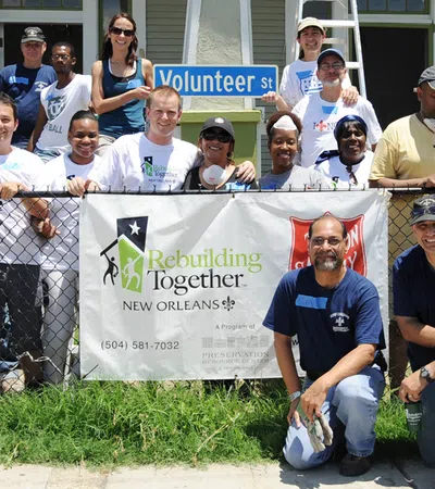 Group of diverse volunteers posing behind a Rebuilding Together New Orleans banner on a sunny day