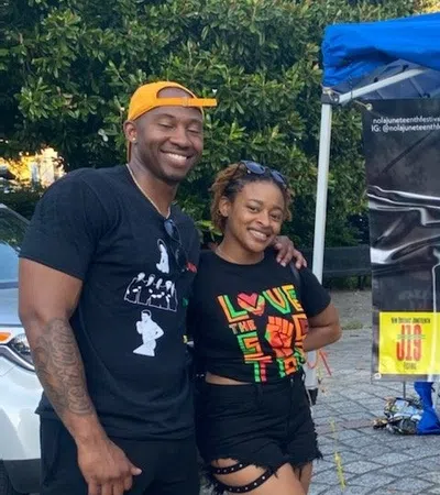 Smiling couple posing in front of a colorful NOLA Juneteenth Festival banner under a blue canopy on a cobblestone path.