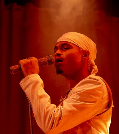 Male singer wearing a white headscarf and jacket performing with a microphone on stage under warm red lighting.