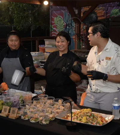 Four chefs preparing and serving a variety of dishes at an outdoor food stall under a wooden roof.