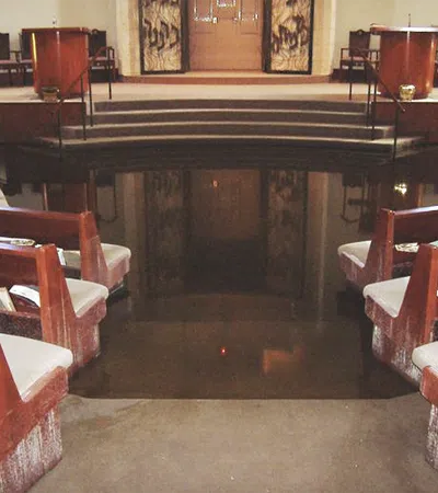Interior of a synagogue with wooden benches, chairs, and a flooded floor reflecting the surroundings.