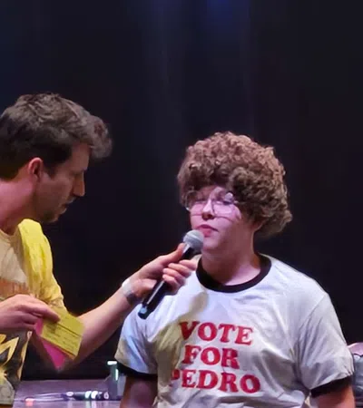 Two men on stage during a performance, one interviewing the other wearing a Vote for Pedro shirt and curly wig.