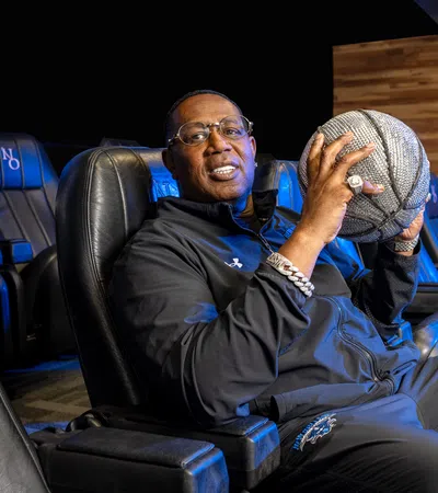 Man in black tracksuit holding a basketball sitting in a New Orleans Basketball lounge area with logo on wall.