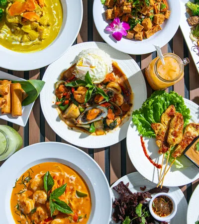 Overhead view of various colorful Thai dishes including curry, stir-fry, spring rolls, and satay with dipping sauces.