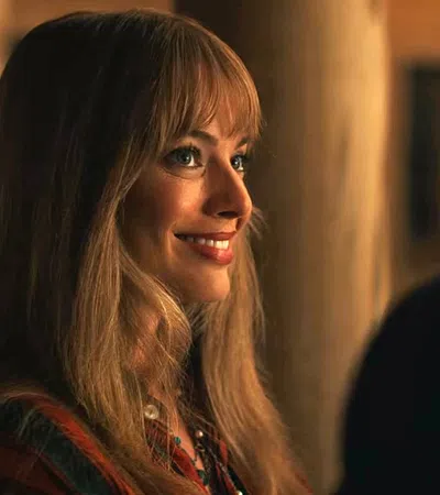 Smiling woman with long blonde hair and bangs looking to the side in warm indoor lighting.
