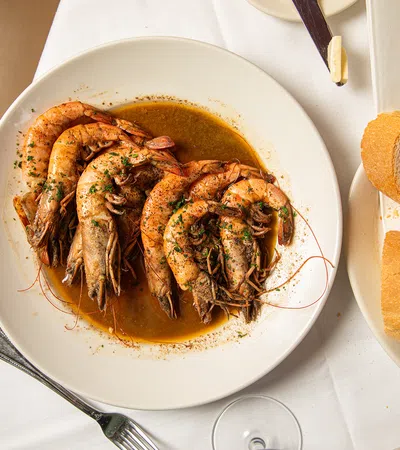 Plate of cooked shrimp in seasoned sauce served with slices of crusty bread on a white tablecloth.