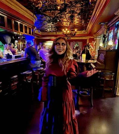 Woman in a red gothic dress with vampire makeup in a dimly lit ornate bar with patrons and warm chandeliers.