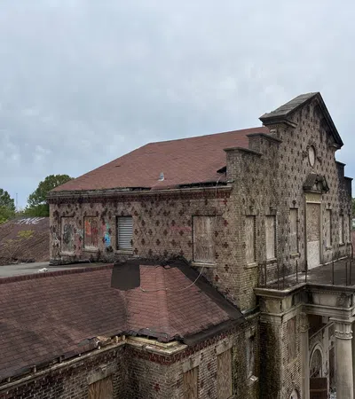 Abandoned brick building with boarded windows, red roof, and classical columns under a cloudy sky.