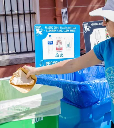 Woman in hat disposing of food waste into a compost bin next to recycling bins at an outdoor event.