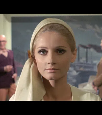 Close-up of a woman with makeup and headscarf, two men blurred in background inside a decorated room.