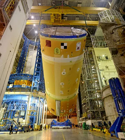 Large orange spacecraft fuel tank suspended inside a spacious industrial assembly facility with workers and machinery.