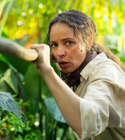 Woman in beige shirt cautiously holding a wooden stick amidst dense green jungle foliage.