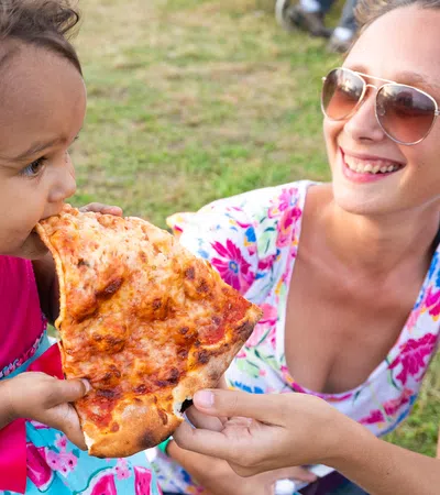 Young child wearing pink dress eating a large slice of pizza outdoors held by a smiling woman in sunglasses.