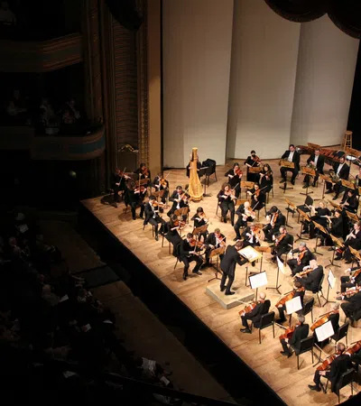 Full orchestra performing on stage in a concert hall with conductor leading musicians and audience seated in darkness