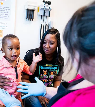 Child in pink shirt interacting with healthcare worker wearing gloves, accompanied by two women in a medical office.