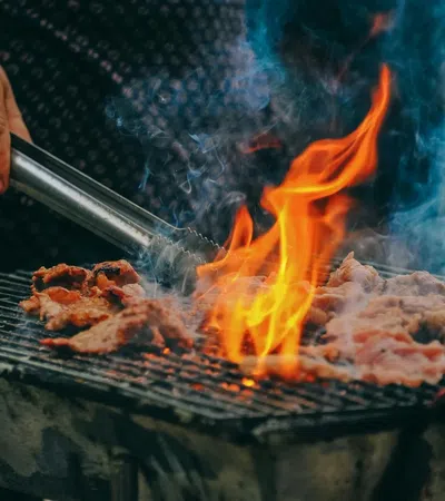 Hand grilling meat over an open flame with smoke rising from a charcoal barbeque grill outdoors.