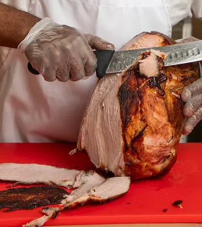 Chef slicing a large smoked ham on a red cutting board wearing gloves and white apron.