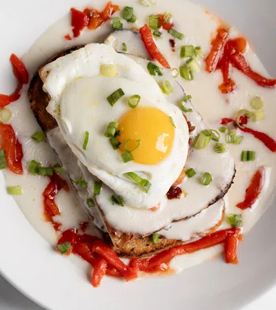 Fried egg atop toast with creamy sauce, red pepper strips, and chopped green onions on a white plate.
