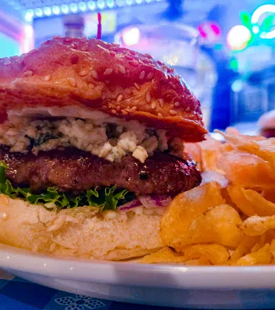 Juicy cheeseburger with lettuce and blue cheese on a sesame bun, served with crispy potato chips on a white plate.