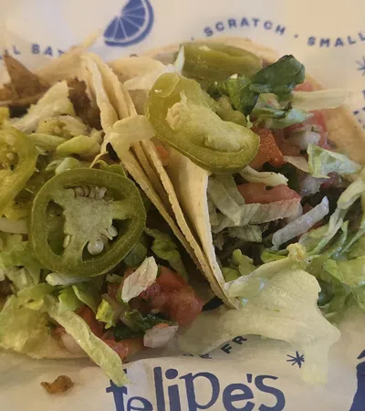 Two soft tacos filled with lettuce, jalapenos, tomatoes, and seasoned meat on branded paper wrapper.