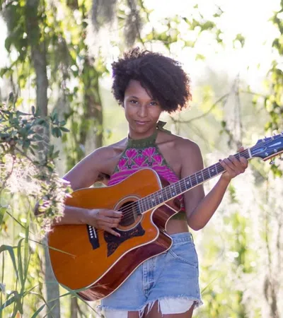 Young woman playing acoustic guitar barefoot in a sunny, lush forest clearing with hanging moss and sand.