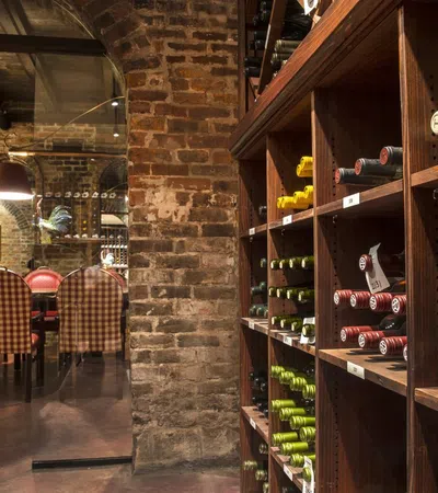 Cozy wine cellar with wooden racks filled with bottles, exposed brick walls, and a dining area with checked chairs.