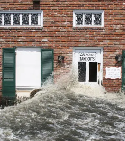 Floodwaters surge against a brick building facade with closed windows and a takeout restaurant door.