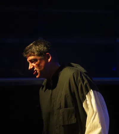 Man in dramatic lighting wearing black shirt with white sleeves, performing or speaking intensely on stage