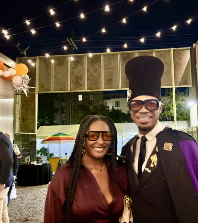 Two stylish people smiling at an outdoor nighttime event with decorative lights and party decorations.
