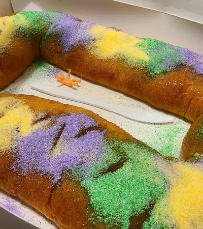 Traditional King Cake decorated with purple, green, and gold sugar in a white box with a plastic knife