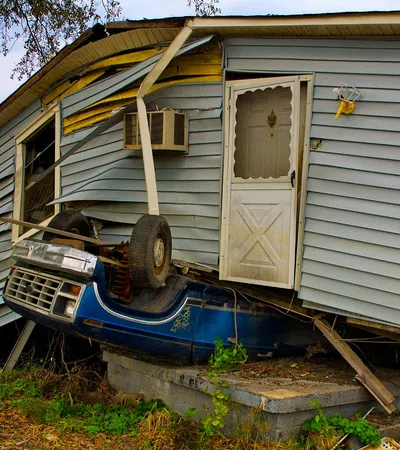A damaged house with the front lifted by a blue car crushing the foundation amid debris and overgrown grass.