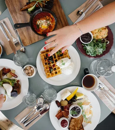 Overhead view of a breakfast table with waffles, eggs, coffee, salad, and people reaching for food.