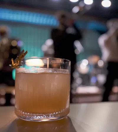 Cocktail glass with an orange drink and garnish on a table in a dimly lit jazz bar with musicians playing in the background