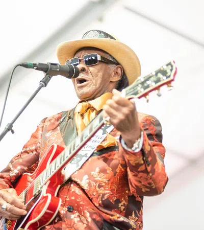 Musician in floral suit and hat playing electric guitar and singing into microphone on stage.