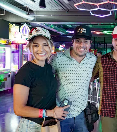 Four friends smiling in a vibrant arcade with colorful neon lights and casual attire including hats and wristbands.